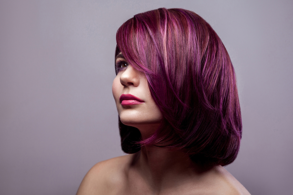 Woman looks off to the side with bare shoulders wearing a dark pinkish purple hair color in front of a gray wall