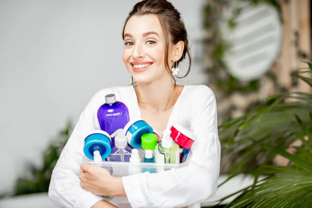 Brunette woman smiles while holding a representation of the top hair products for thin hair in her arms with multi-colored bottles and jars
