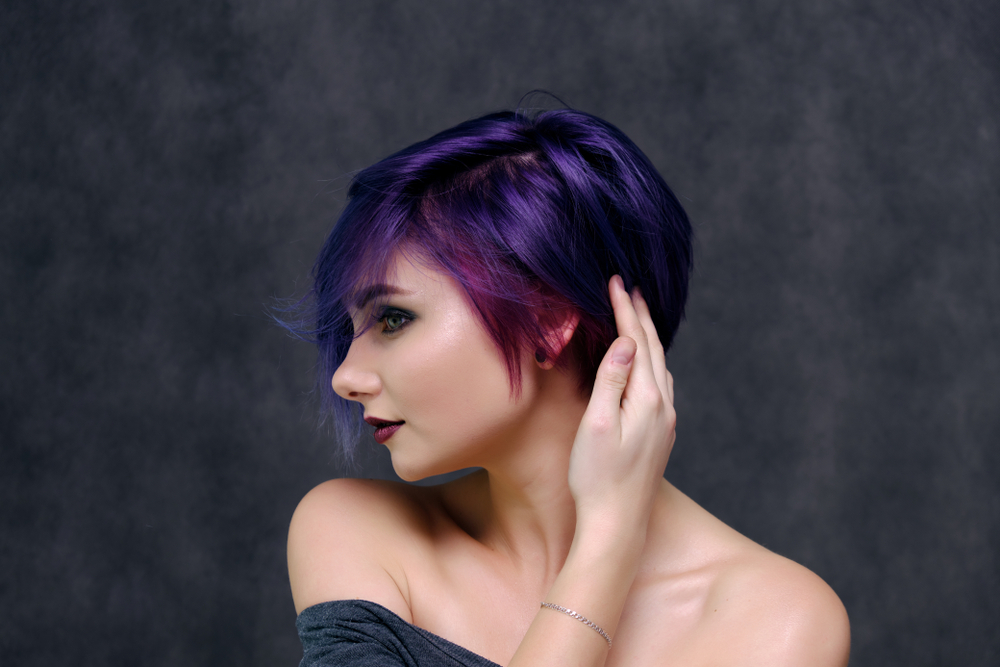 Woman looks over to the side with her hand touching her dark purple hair with pinkish accents in front of gray backdrop