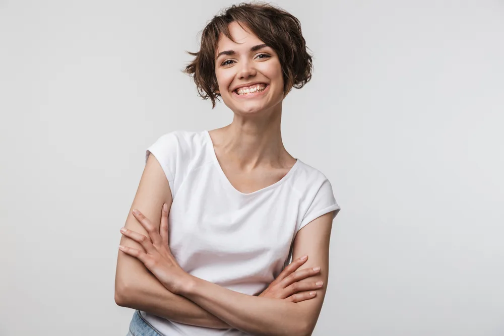 Smiling brunette woman with white t-shirt against white background crosses her arms with a short bob and very wispy bangs