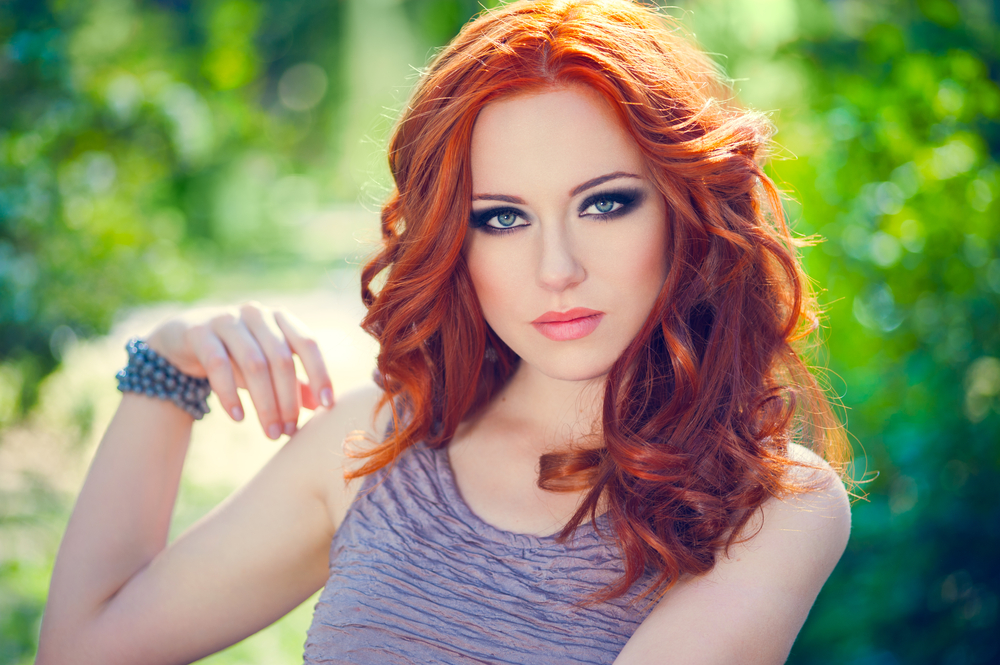 Woman outdoors with glam eye makeup gazes at camera with one of the top warm red hair colors in an auburn with copper highlights shade