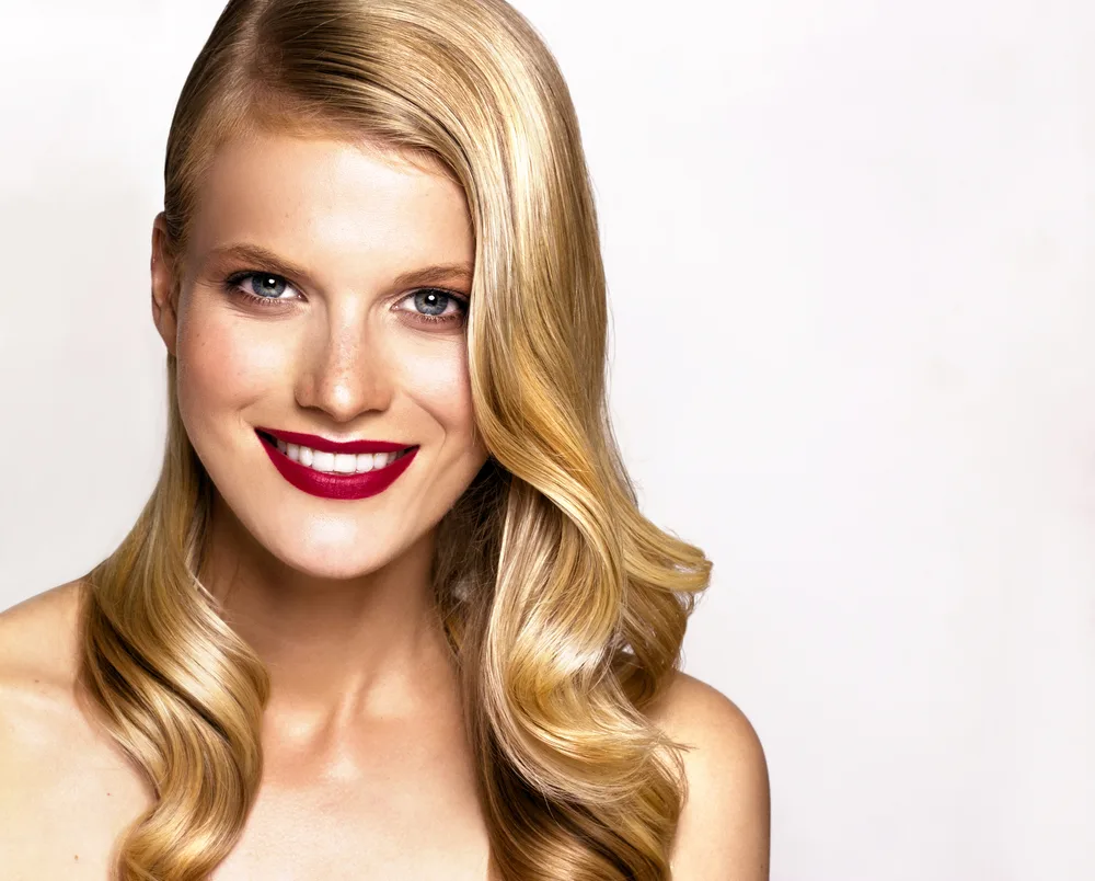 Woman smiles wearing red lipstick and honey blonde color in hair with bare shoulders on white background