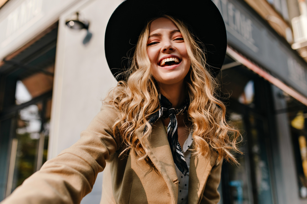 Young woman with curly honey blonde hair color wears trendy hat and coat smiling in the city