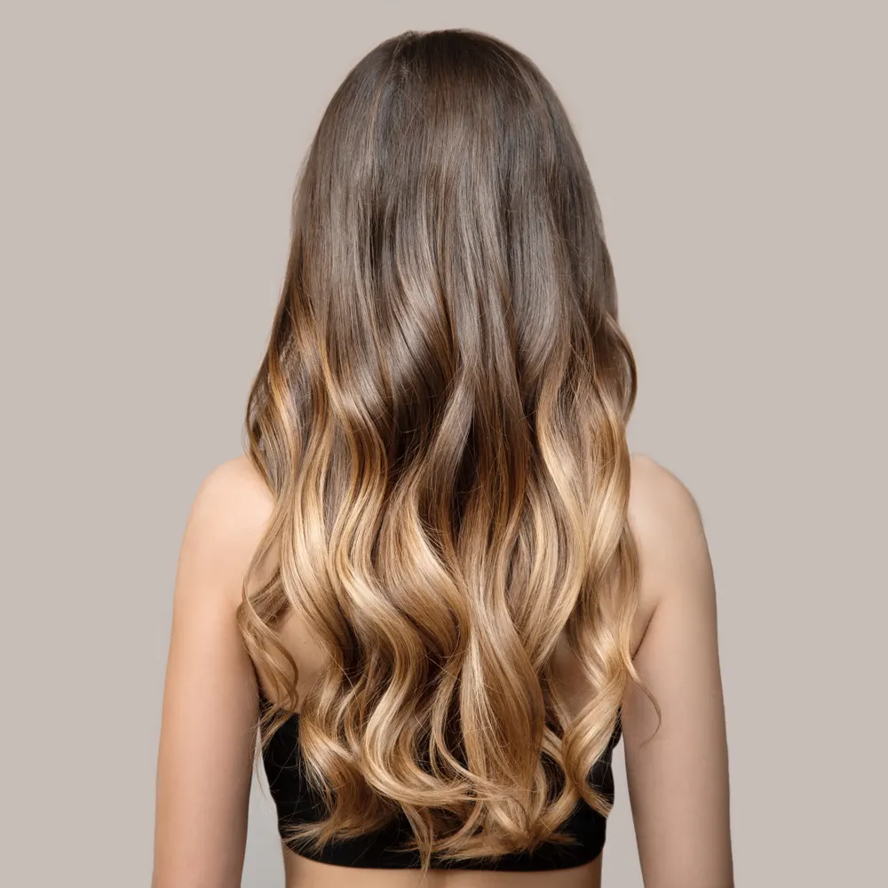 Brunette woman shown from back view with blonde balayage color shows off medium beach waves hairstyle in front of gray wall