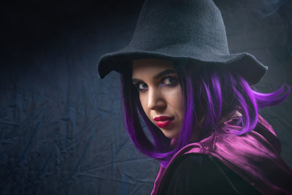 Woman with purple hair dressed up as a witch for Halloween wearing black hat and looking over her shoulder