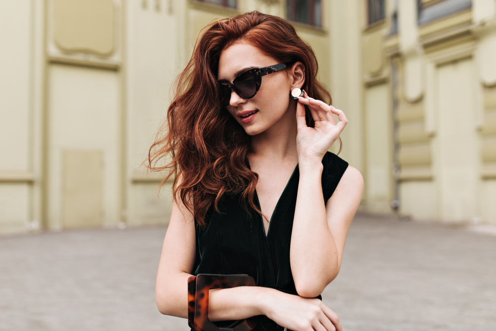 Woman in dark sunglasses holding purse adjusts earring with dark auburn warm red hair color