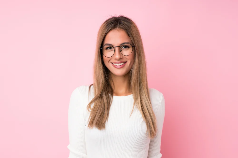 Smiling woman with glasses stands in front of pink wall with dark golden blonde hair to showcase the best hair color for age reversal