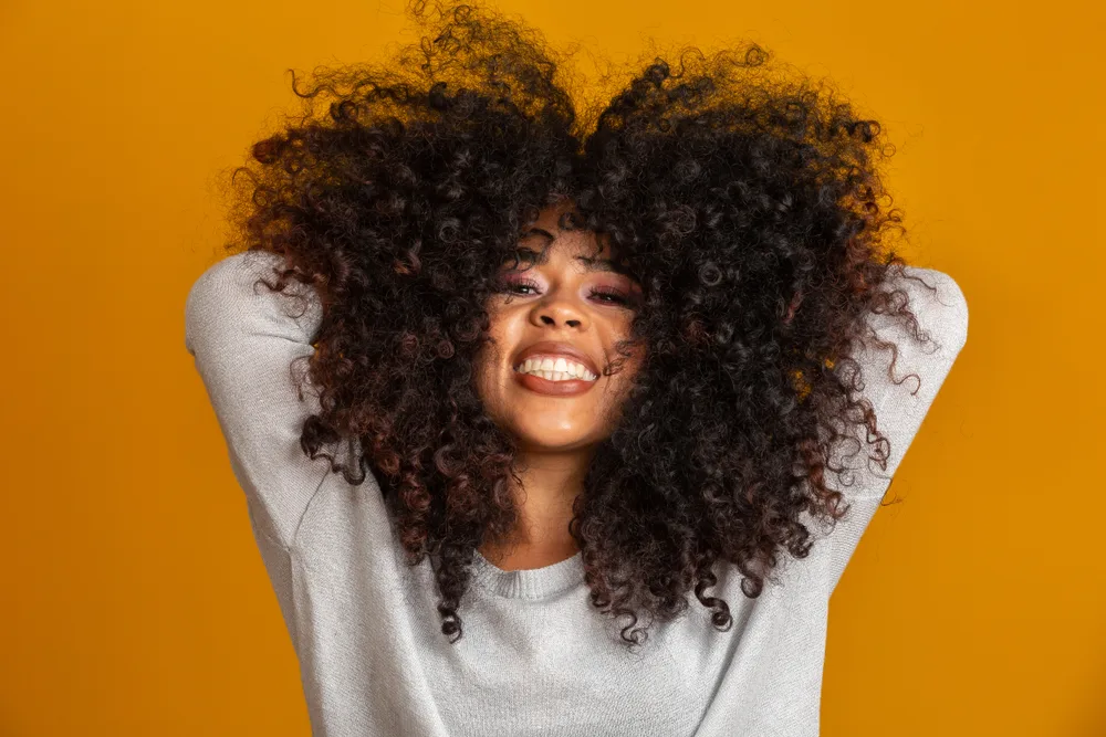 Black woman holds hands behind her head in front of orange wall and shows off her healthy hair texture that can be achieved using black owned hair products