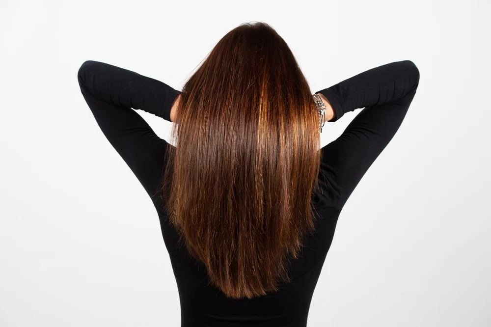 Woman shown from rear with arms raised to lift her long dark brown hair with copper highlights wearing black long sleeved shirt
