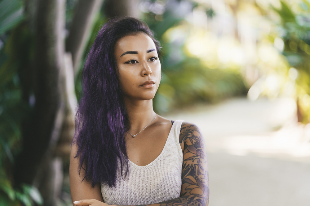 Woman with arm tattoos and white tank top crosses her arms looking off into the distance outdoors with dark purple hair color