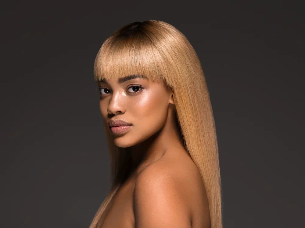 African American woman glances at camera with bare shoulders in front of dark gray wall with honey blonde hair color, straight texture, and dark brown roots