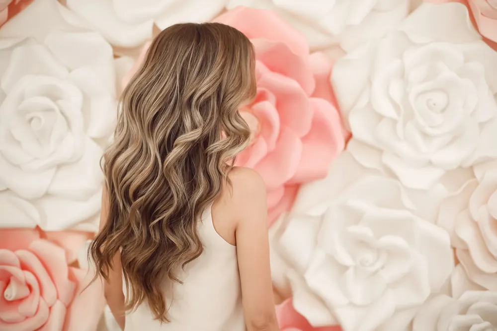 Back view of woman with blonde balayage in front of floral wall showing off medium beach waves hair style in a glamour look