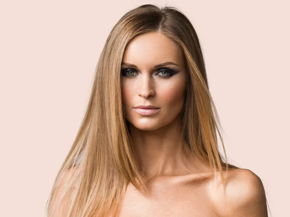 Serious woman with straight honey blonde hair stares directly at camera with bare shoulders in front of light peach background
