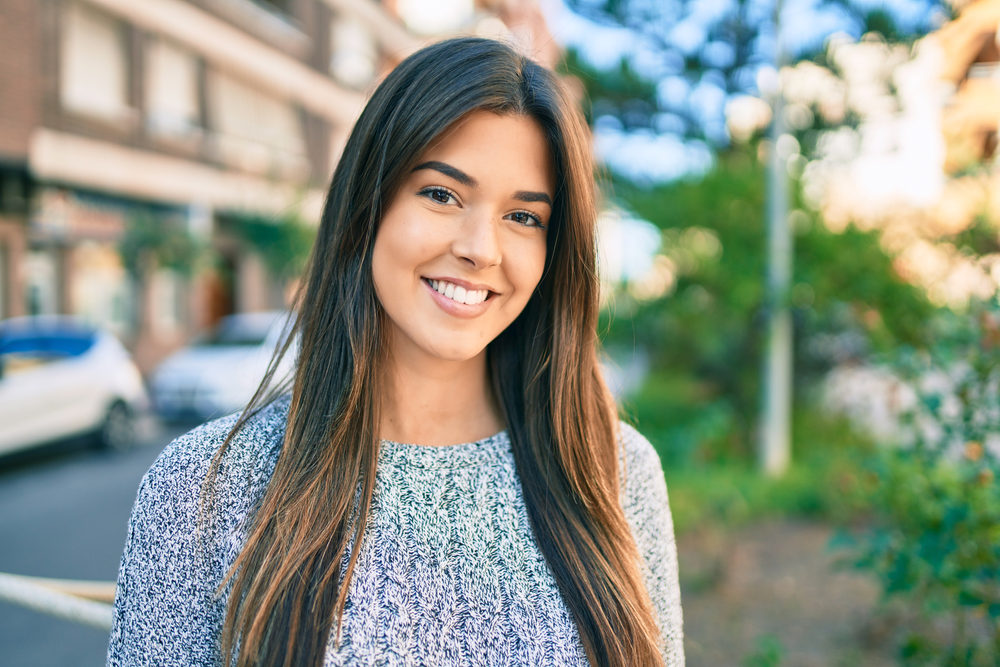 Smiling woman on a city street wears a gray sweater and straight long dark brown hair with golden brown highlights