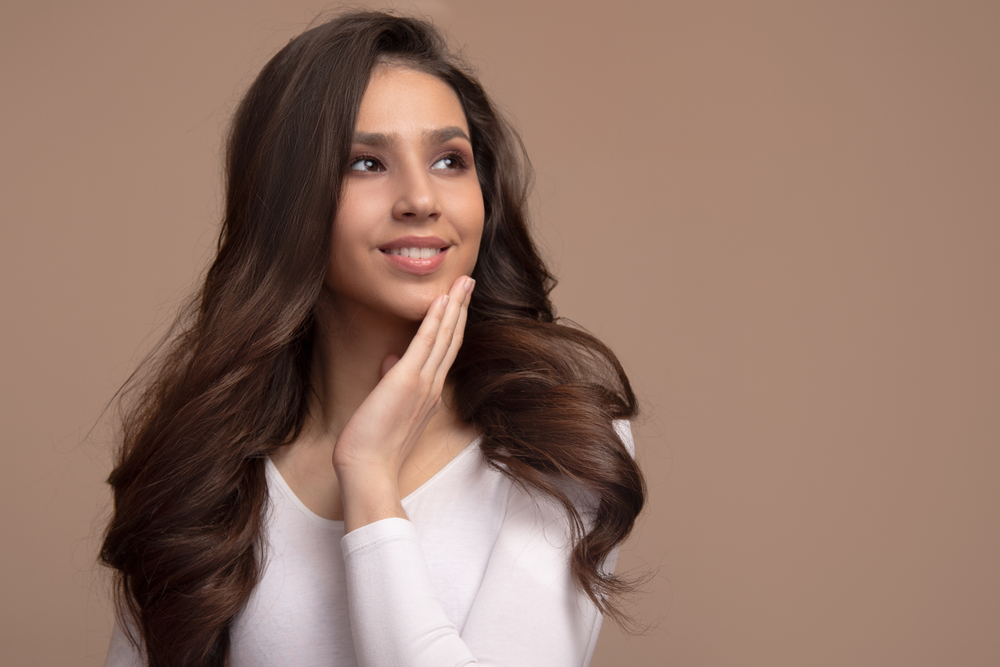 Brunette woman with long thin hair styled in a voluminous blowout poses with her hand placed on her chin in front of a light brown background in a white v-neck shirt