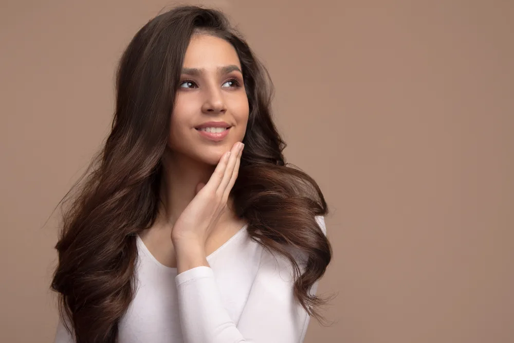 Brunette woman with long thin hair styled in a voluminous blowout poses with her hand placed on her chin in front of a light brown background in a white v-neck shirt