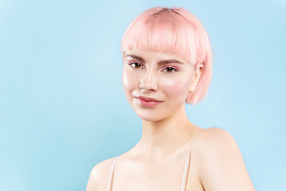 Woman with glowing skin and light pink hair wears a tank top in front of a blue background with short micro bangs hairstyle with blunt bob