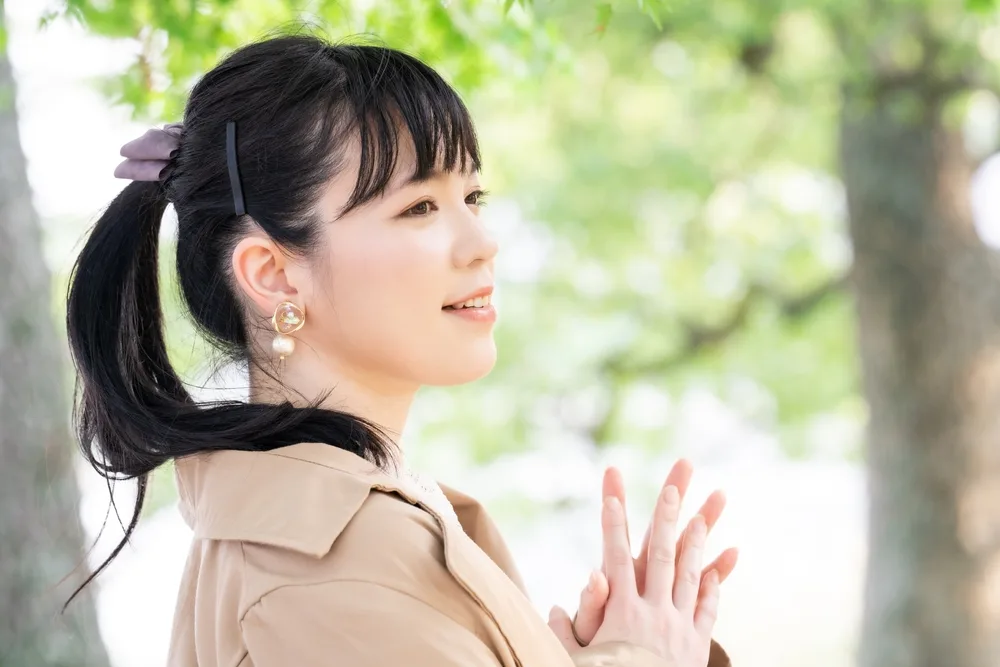 Dark-haired Asian woman touches hands together outdoors with hair in a ponytail and textured wispy fringe