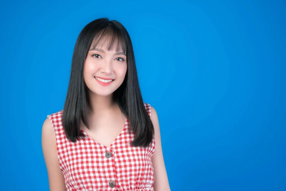 Smiling woman with wispy blunt bangs stands in front of blue background wearing a red gingham dress
