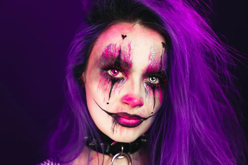 Woman dressed up in one of several Halloween costumes for purple hair with clown makeup and colored contact lenses