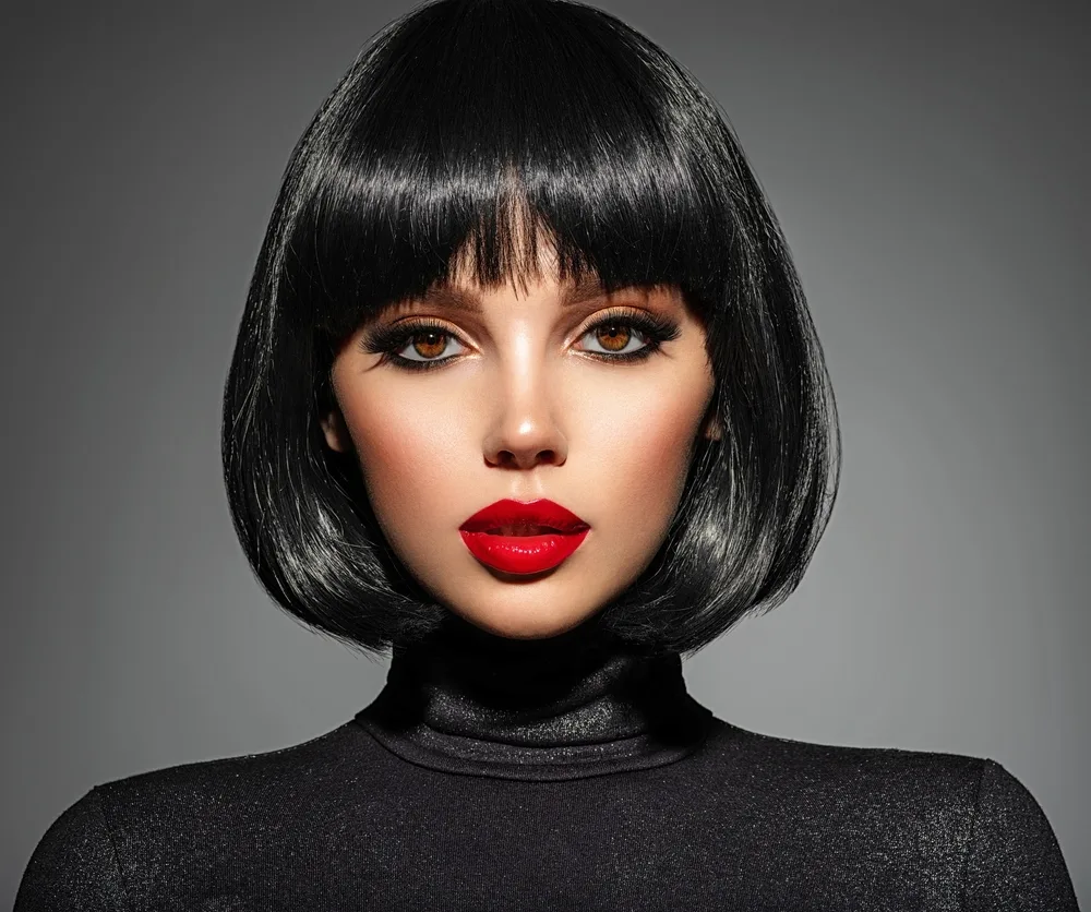 Woman with black bob and bangs hairstyle wears a black turtleneck in front of gray background with red lipstick