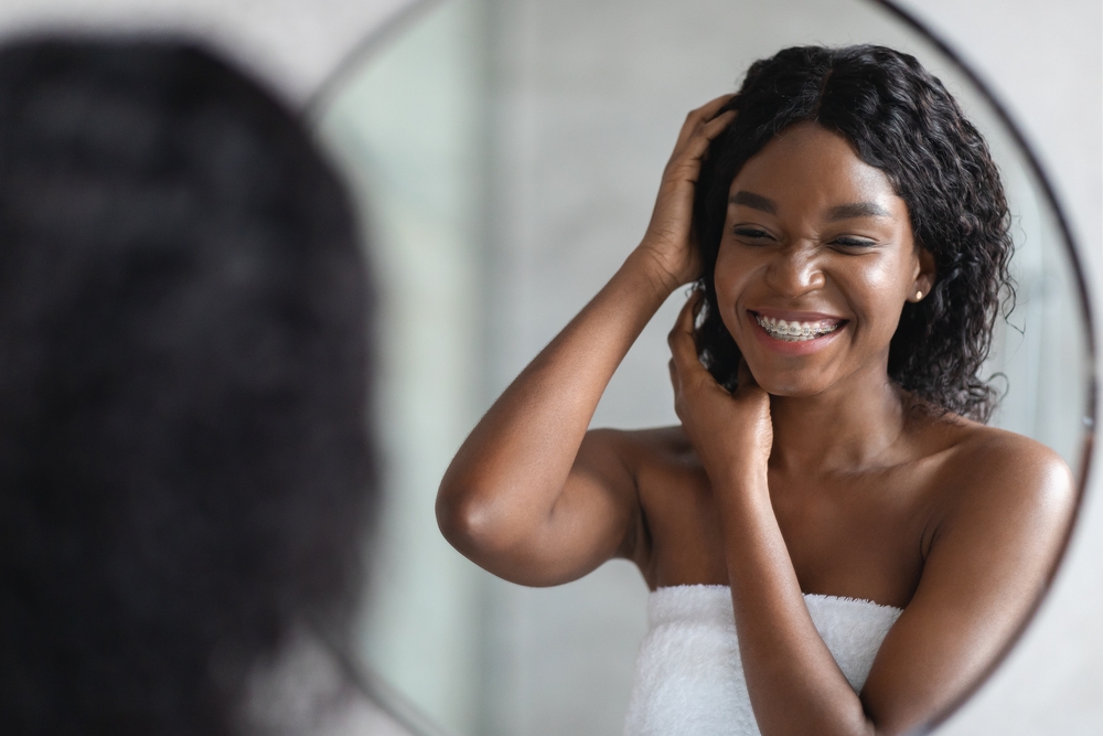 Black woman smiles in mirror while styling her freshly washed coily hair with products made by black owned businesses