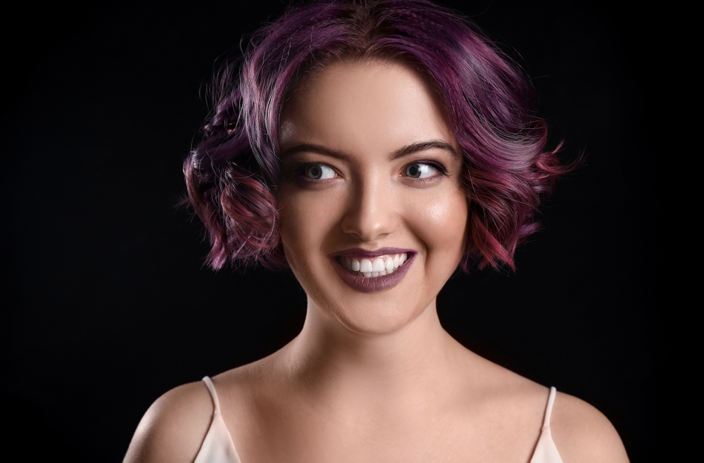 Woman looks away and smiles wearing a spaghetti strap tank top with dark purple and maroon hair color
