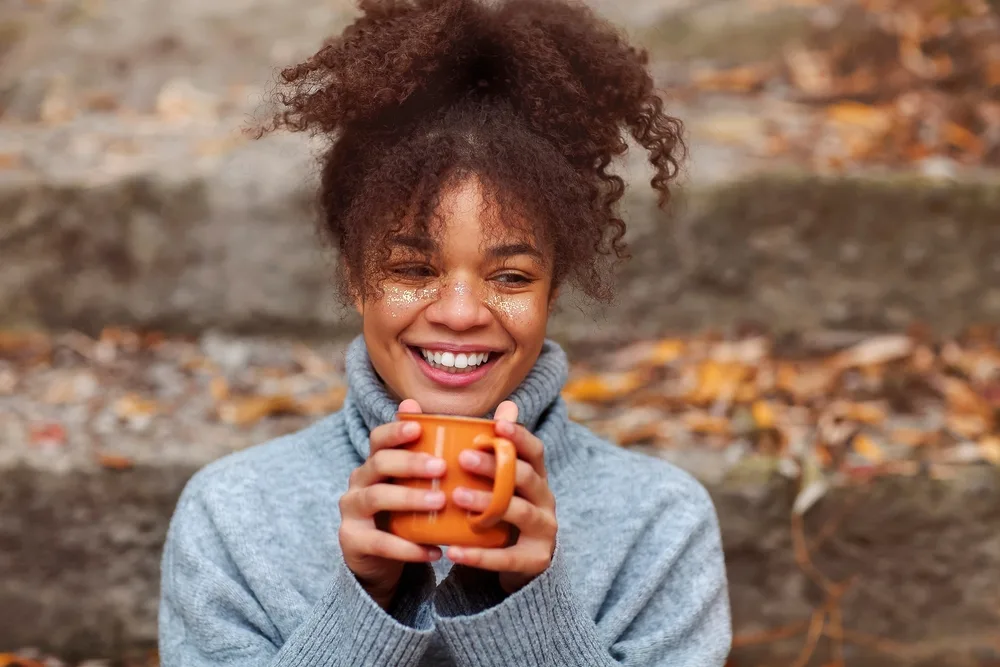 Cheerful woman with brown skin holds an orange mug and smiles with her coily hair in a ponytail and curly bangs cut in a wispy style