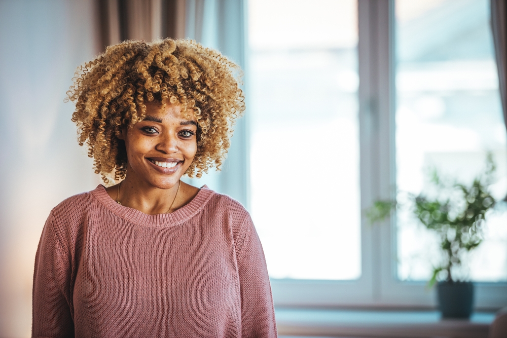 Black woman smiles wearing pink sweater in front of window with honey blonde highlights and coily hair
