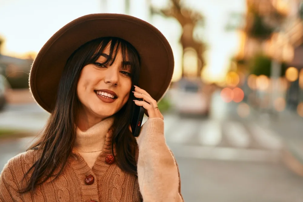 Brunette woman with straight long hair and wispy bangs smiles wearing hat and touching her cheek