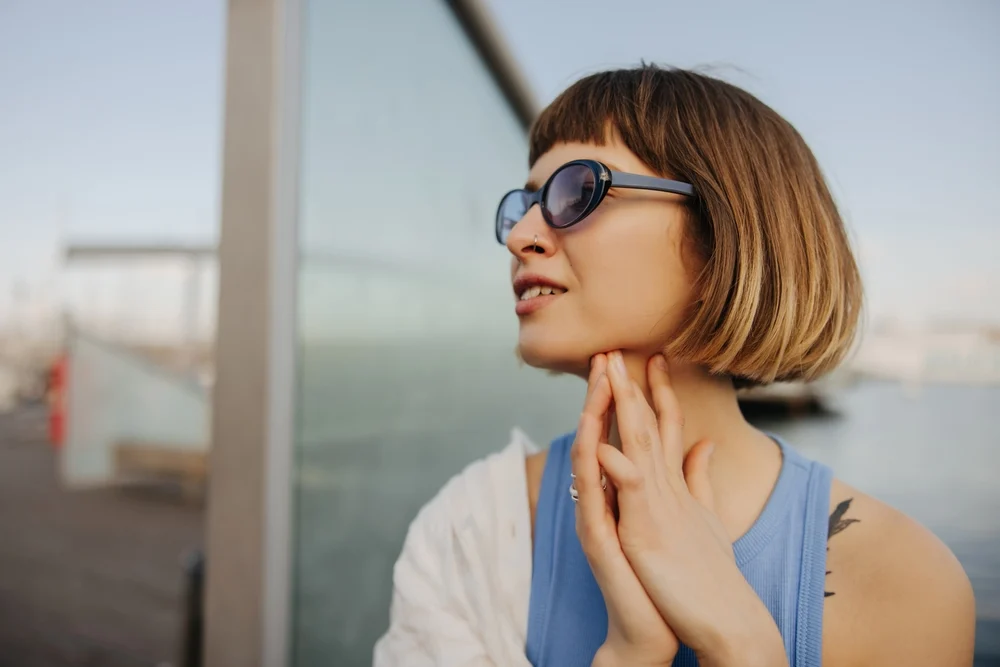 Woman with sombre short hair color and blue sunglasses looks off into the distance with hands clasped