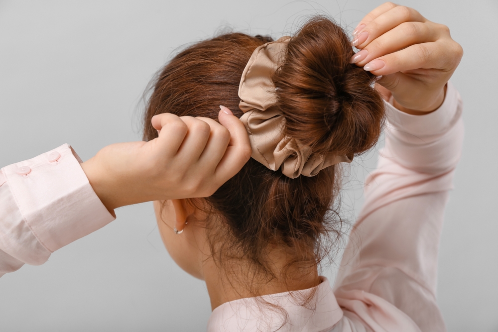 Back view of woman in long sleeves tying her hair into a scrunchy bun as an example of a cute hairstyle for long hair
