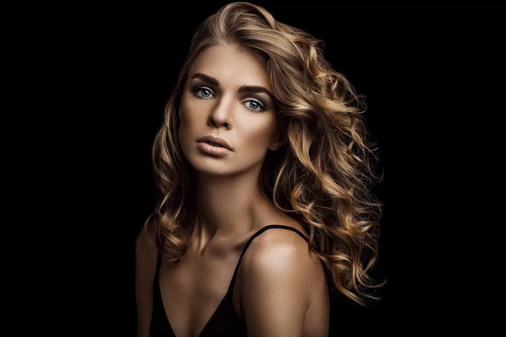 Woman posing in darkness with black tank top has honey blonde hair color with styled curls