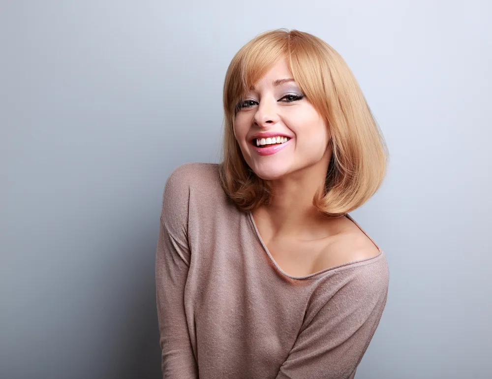 Happy woman with off-shoulder sweater leans forward in front of gray wall with strawberry blonde bob on short hair
