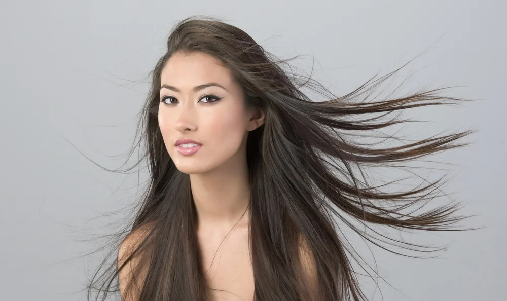 Asian woman glances at camera with her ash-toned dark brown hair blowing in the wind in front of a gray wall