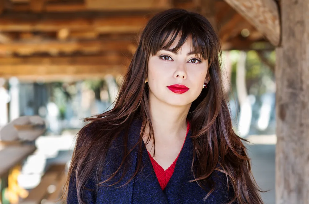 Brunette woman with bold red lips shows off her blunt wispy bangs and long hair wearing v-neckline sweater