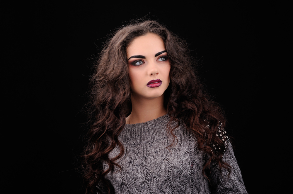 Woman glances off to the side wearing gray cable knit sweater and long curly dark brown hair with mahogany ombre