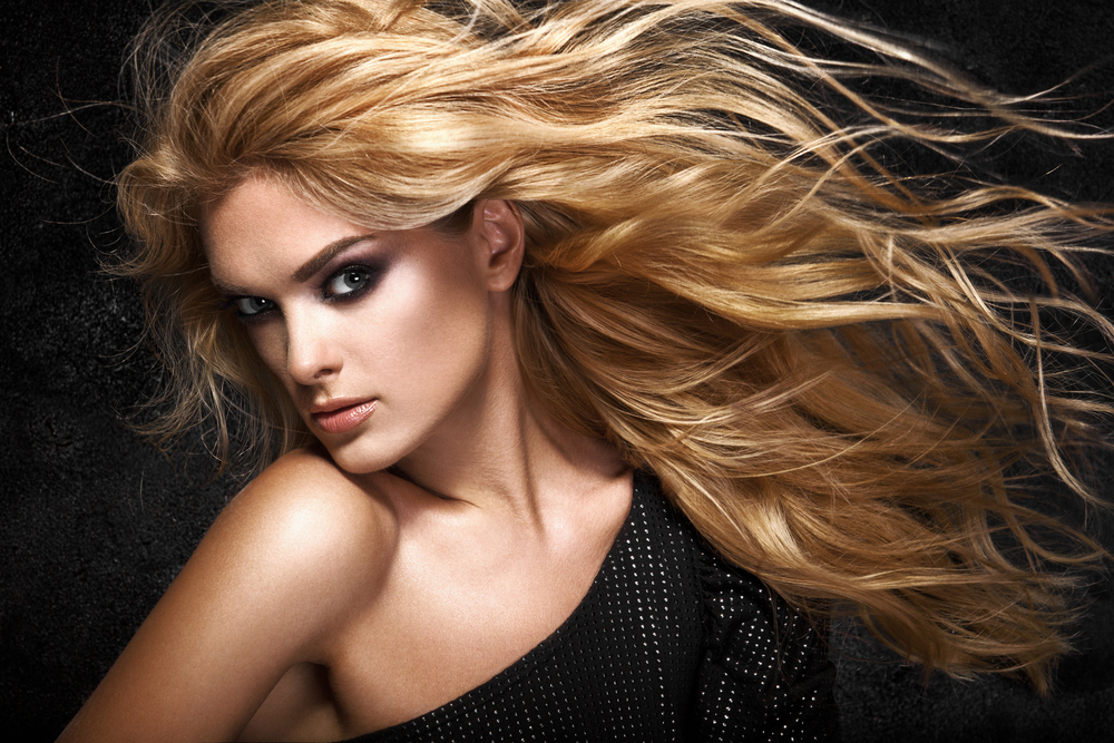 Woman with head turned looks at camera in front of black background with honey blonde hair color and flowing wavy hair