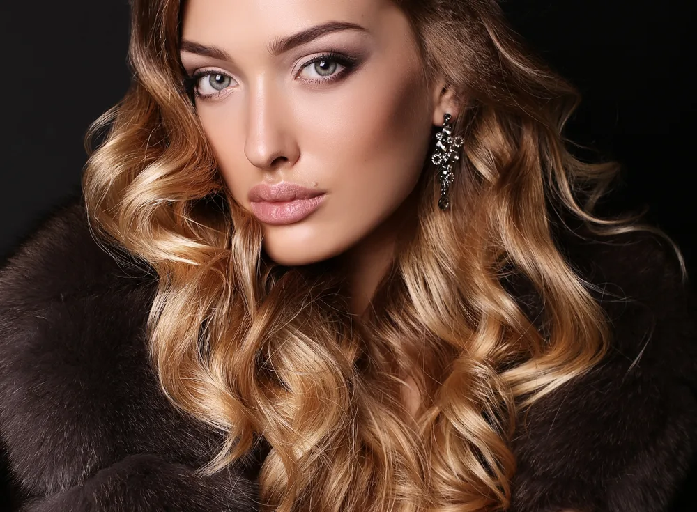 Woman wearing fur coat and dangle earrings stares intensely with honey blonde hair styled in curls