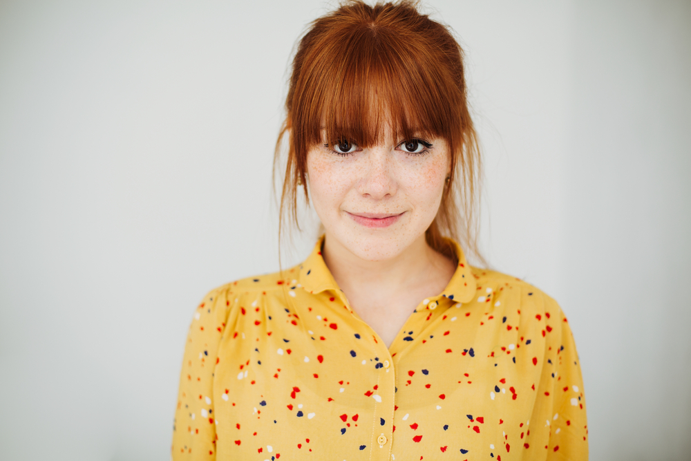 Redheaded woman in patterned yellow shirt slightly smiles wearing a ponytail with thin eyelash length bangs