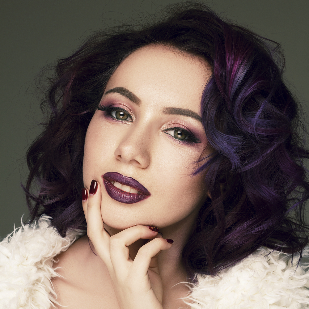 Woman poses with her hand on her chin wearing dark purple hair, lipstick, and eyeshadow