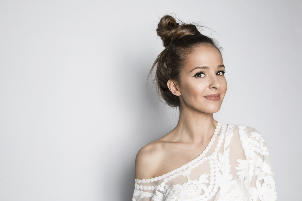 Woman with off-shoulder top stands in white room with one of the cute hairstyles for long hair in a messy top knot
