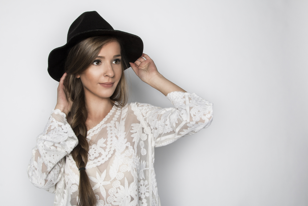 Woman wearing hat and white lace top touches her hair with loose side braid style