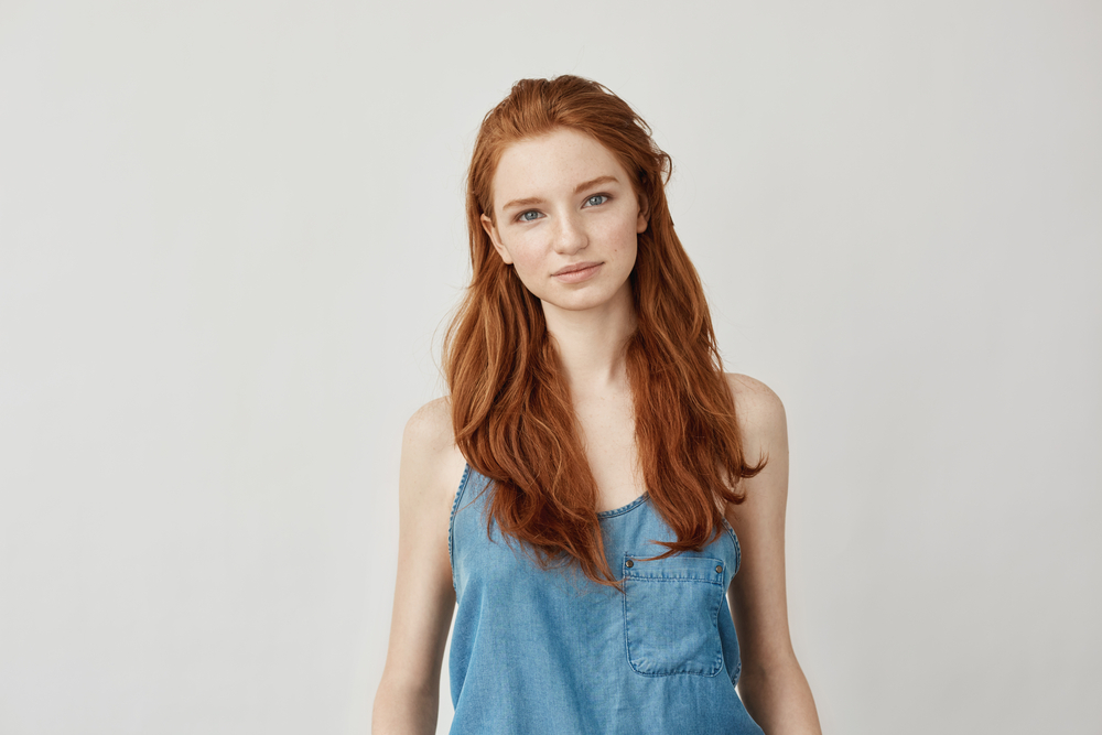 Redhead woman in denim dress slightly smiles against a light background with cinnamon, one of the trendiest warm red hair colors