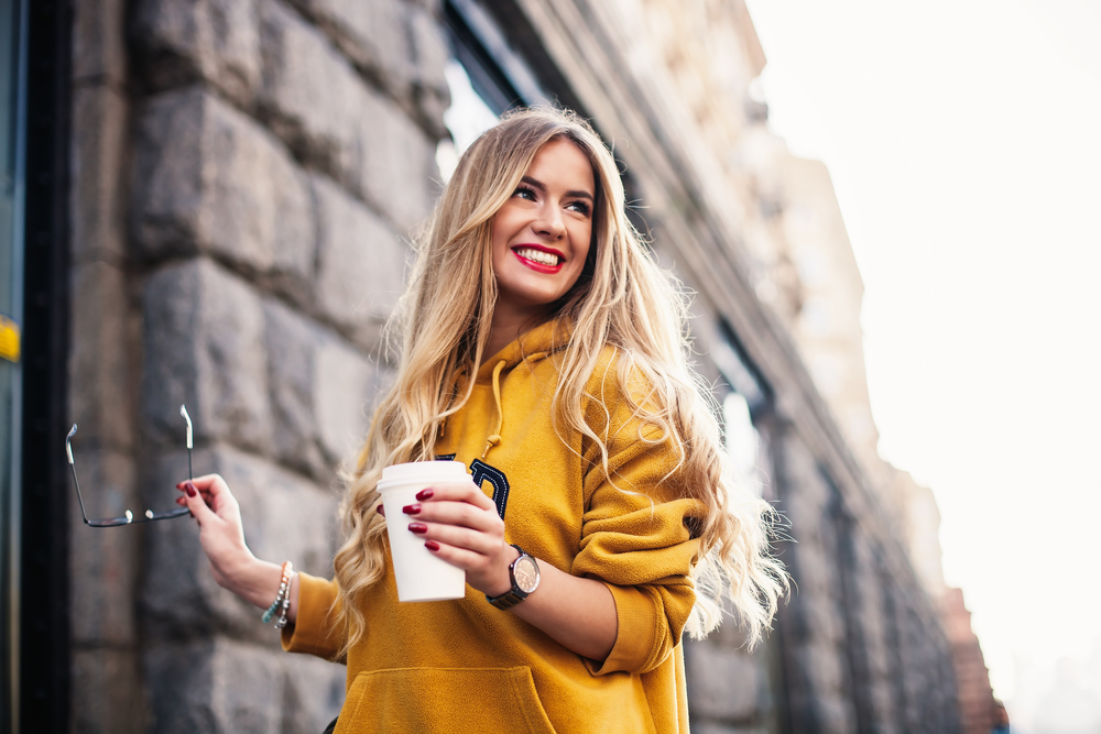 Smiling woman in yellow coat holds coffee cup and glasses while walking in the city with curtain bangs hairstyle