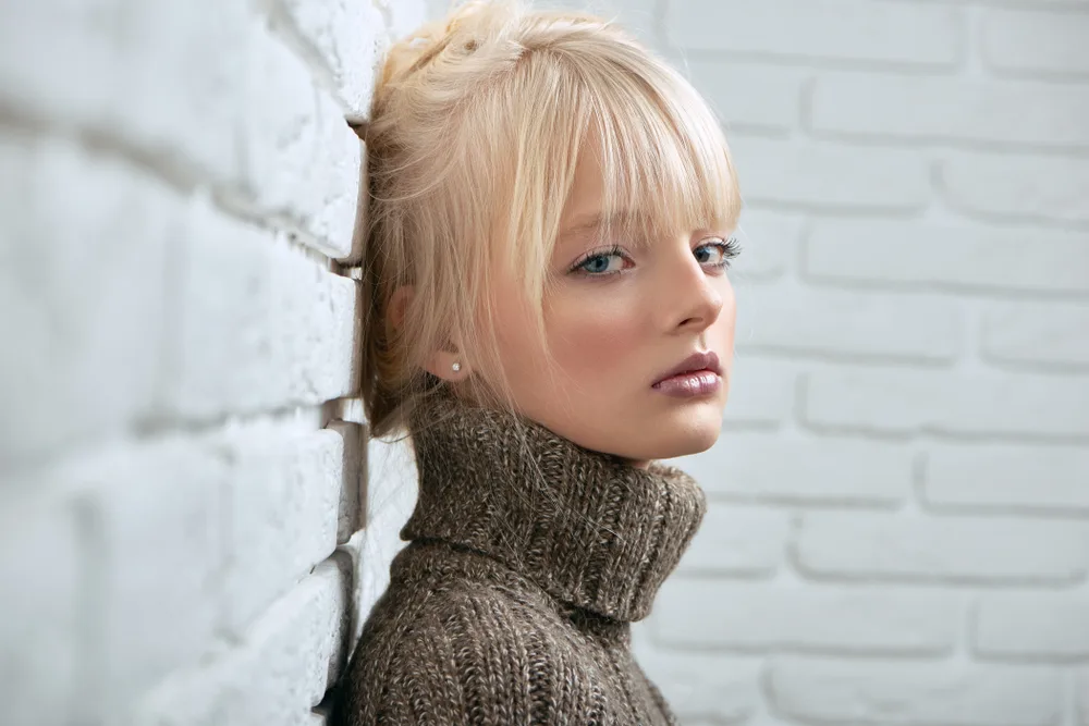 Serious light blonde woman with wispy bangs stands in front of white brick wall with turtleneck sweater on