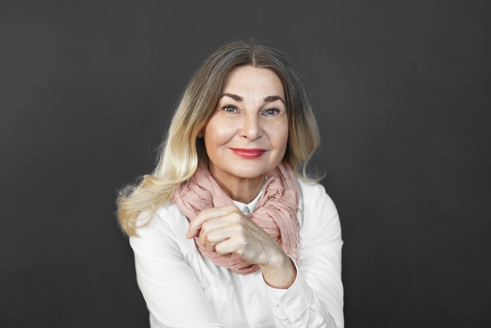 Mature woman on dark background smiles wearing the best hair color for age with a blonde ombre