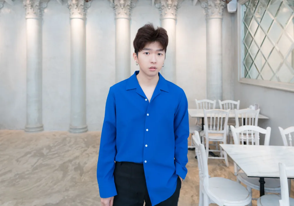 Young Korean man looks at camera in room with white columns wearing a half-tucked blue dress shirt and fluffy two block hairstyle