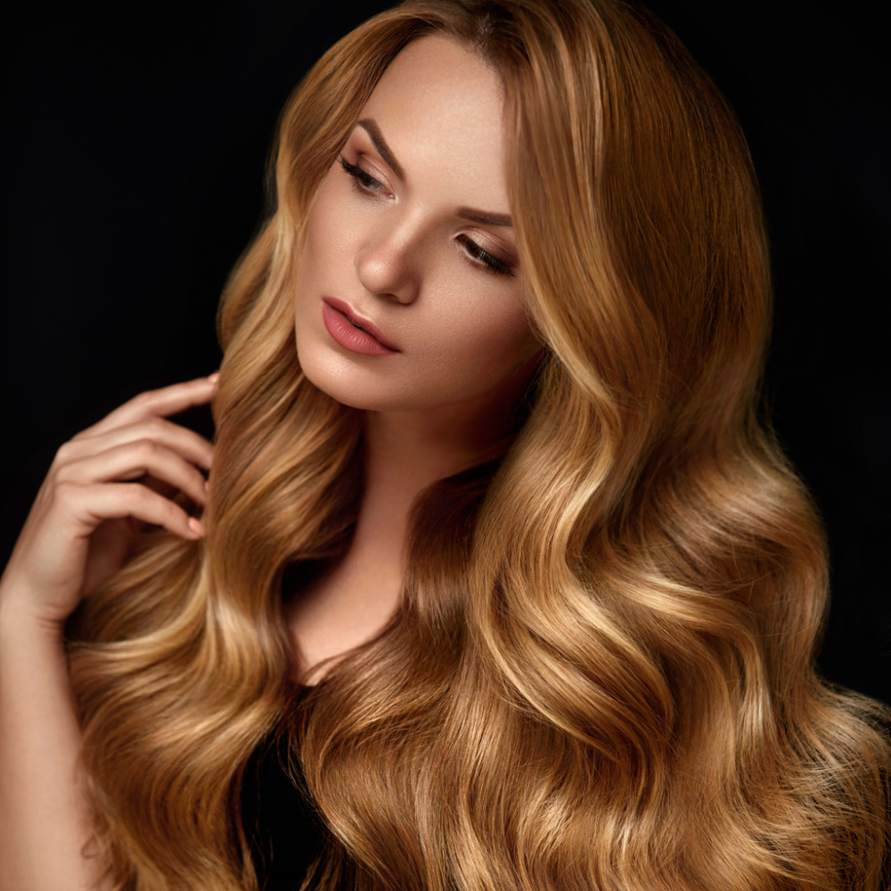 Woman touches her wavy hair in front of black wall showing one of the many shades of blonde in a strawberry golden color
