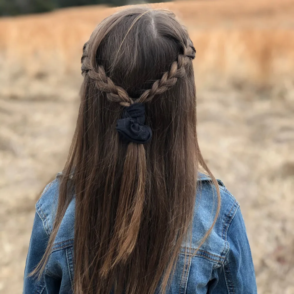 Girl in denim jacket stands in a field seen from behind wearing one of the cutest half up half down hairstyles with a braid